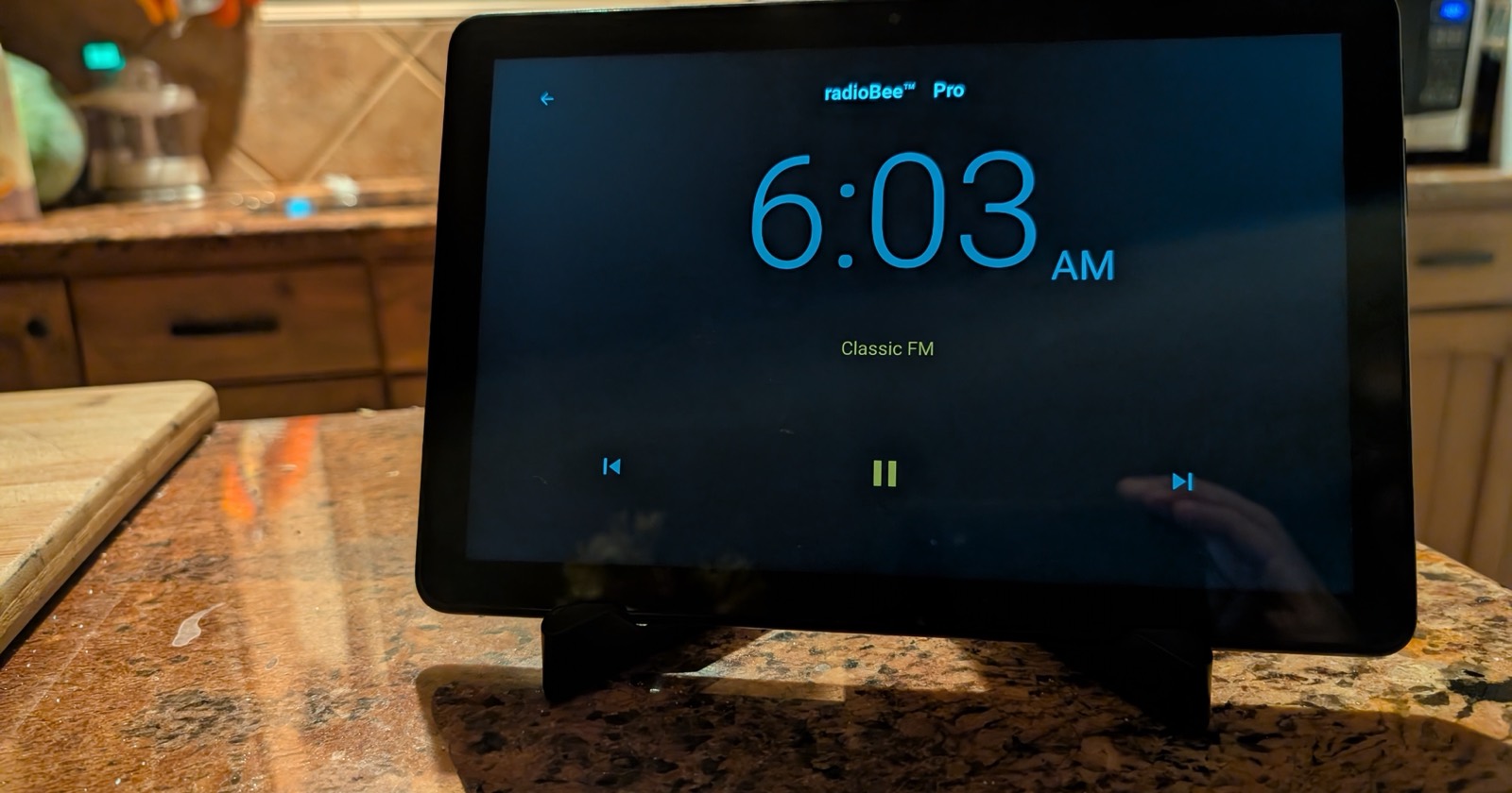Old Fire tablet on a kitchen counter showing radioBee Dock Mode: a large clock, the station name, and playback controls